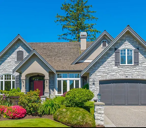 Charming stone house with red front door, gray garage, large windows, lush green lawn, and colorful garden flowers under a clear blue sky — perfect suburban home exterior.